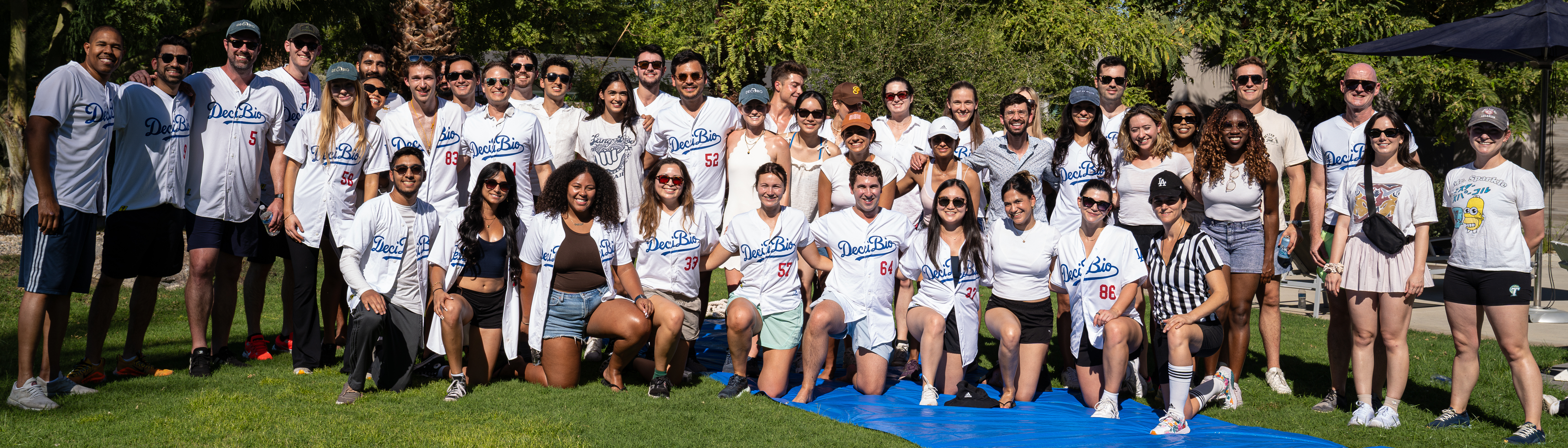 A large group of men and women gathers on a sunny lawn, most wearing white baseball-style jerseys labeled “DeciBio,” while others are dressed in casual warm-weather clothing. They stand and kneel together, smiling at the camera, with trees and greenery in the background.
